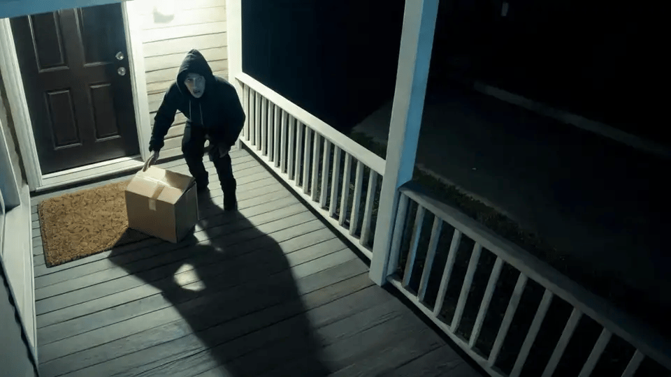 A person in a hoodie reaching for a parcel is caught in a floodlight